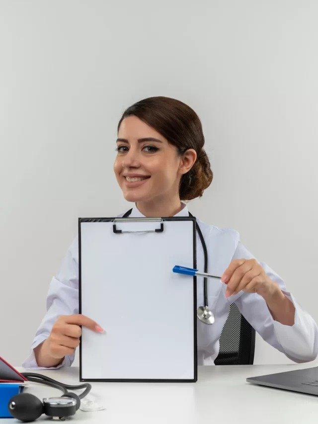 cropped-smiling-young-female-doctor-wearing-medical-robe-stethoscope-sitting-desk-with-medical-tools-laptop-holding-clipboard-pointin